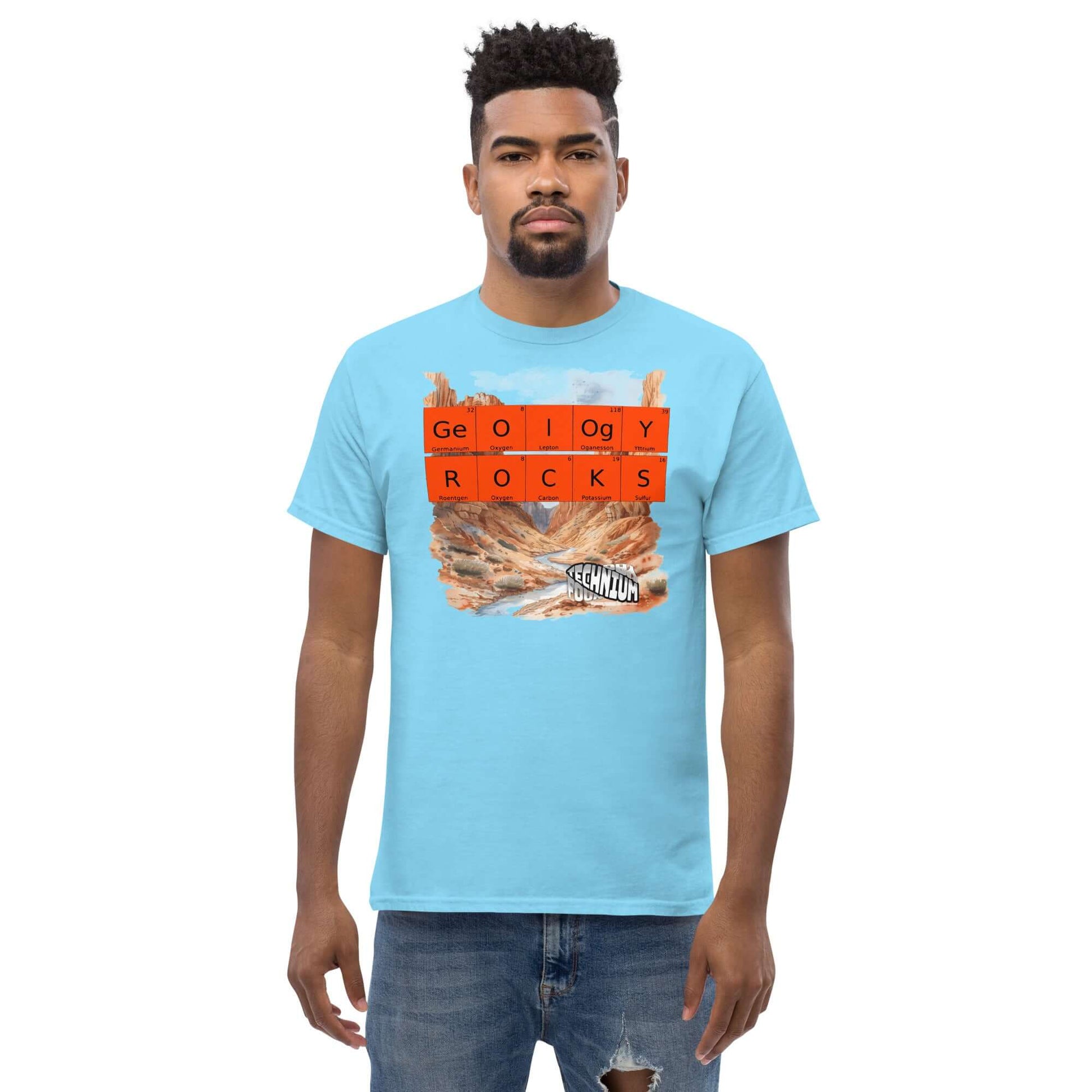 Man wearing a blue "GEOLOGY ROCKS" T-shirt by Technium Foundry with periodic table design and scenic landscape background.