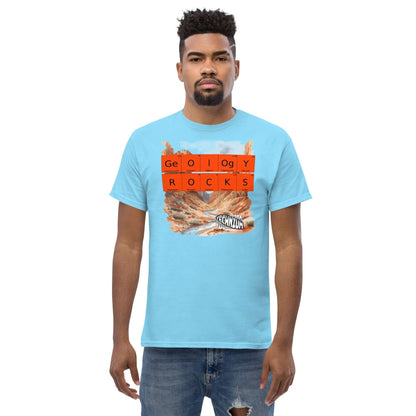 Man wearing a blue "GEOLOGY ROCKS" T-shirt by Technium Foundry with periodic table design and scenic landscape background.