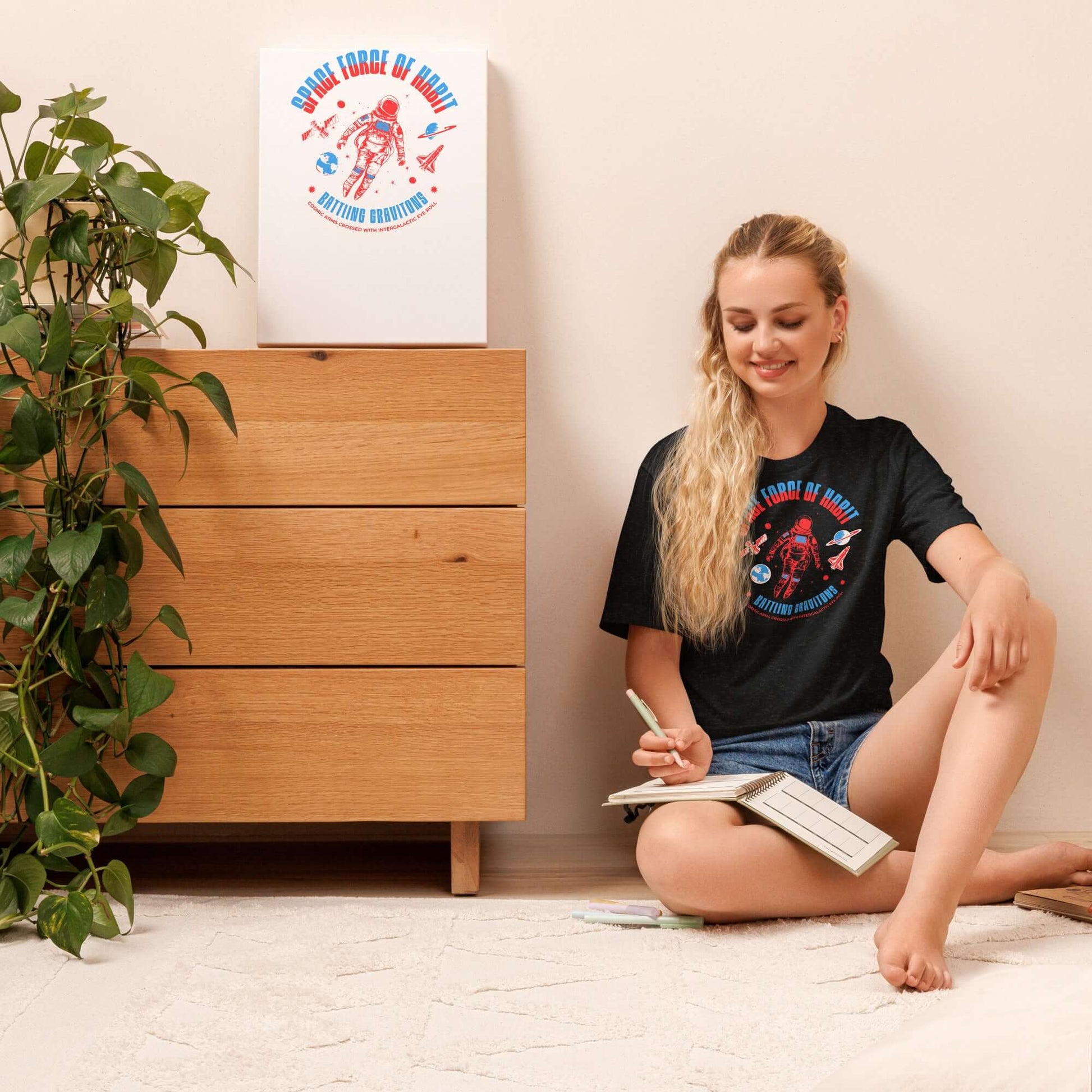 Woman wearing Space Force of Habit T-Shirt featuring astronaut humor design, sitting on floor near dresser and plant.