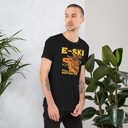 Man wearing black E-SK8 'Till My Battery Dies' T-Shirt featuring electric Grim Reaper design in yellow, standing beside green plants.