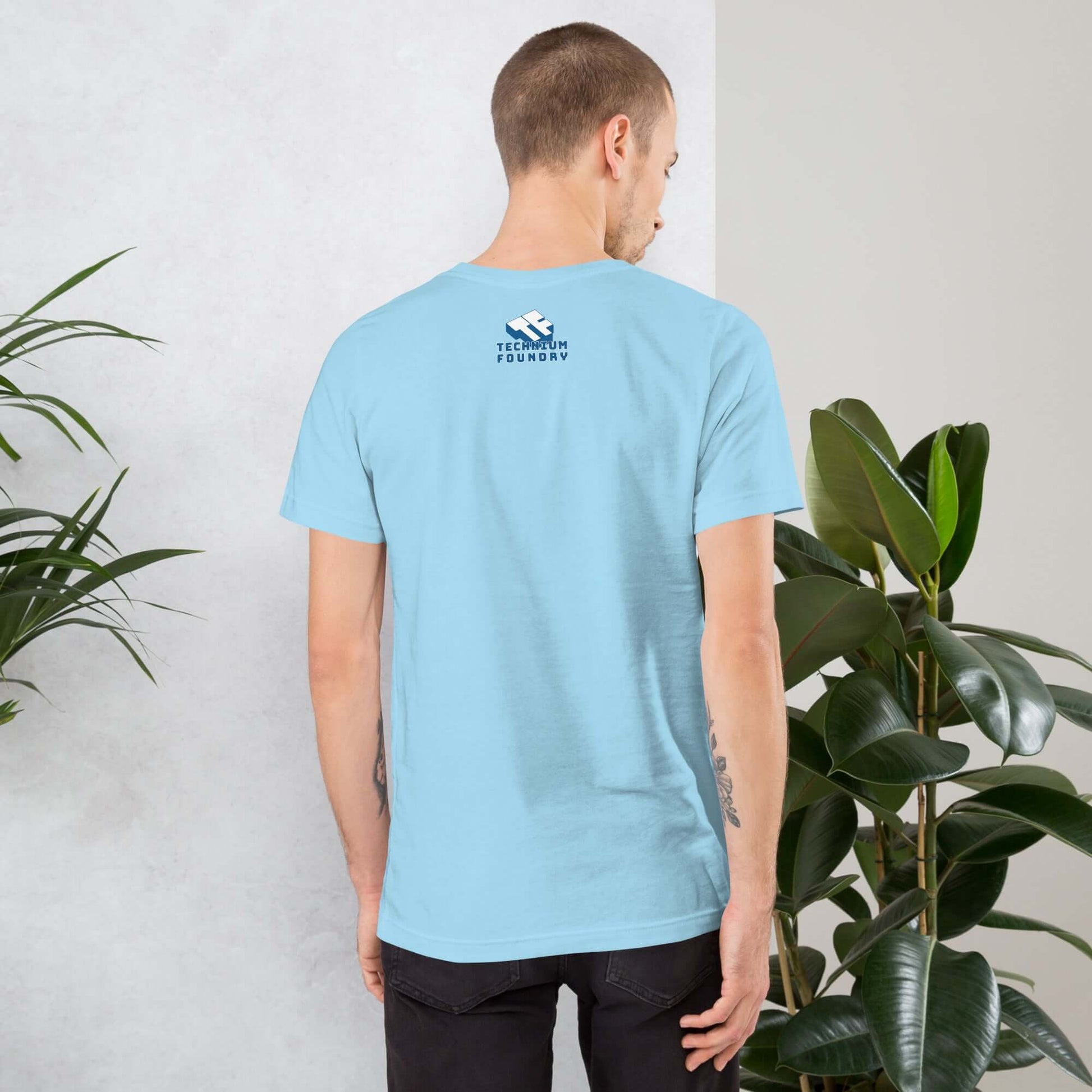 Man wearing light blue T-shirt with logo, standing near plants. Back view.