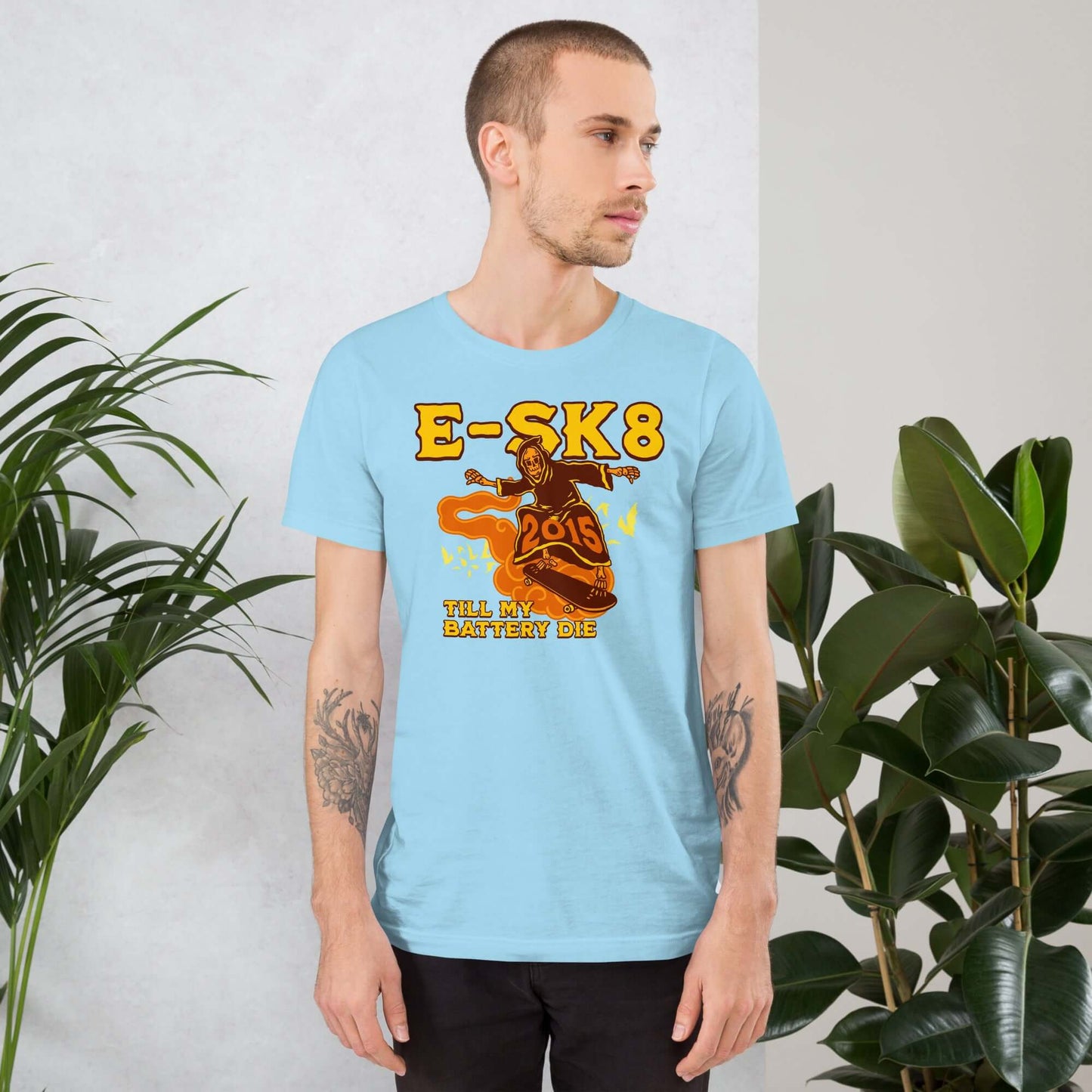 Man wearing 'E-SK8 Till My Battery Dies' Grim Reaper electric revolution t-shirt in blue, standing by green plants.