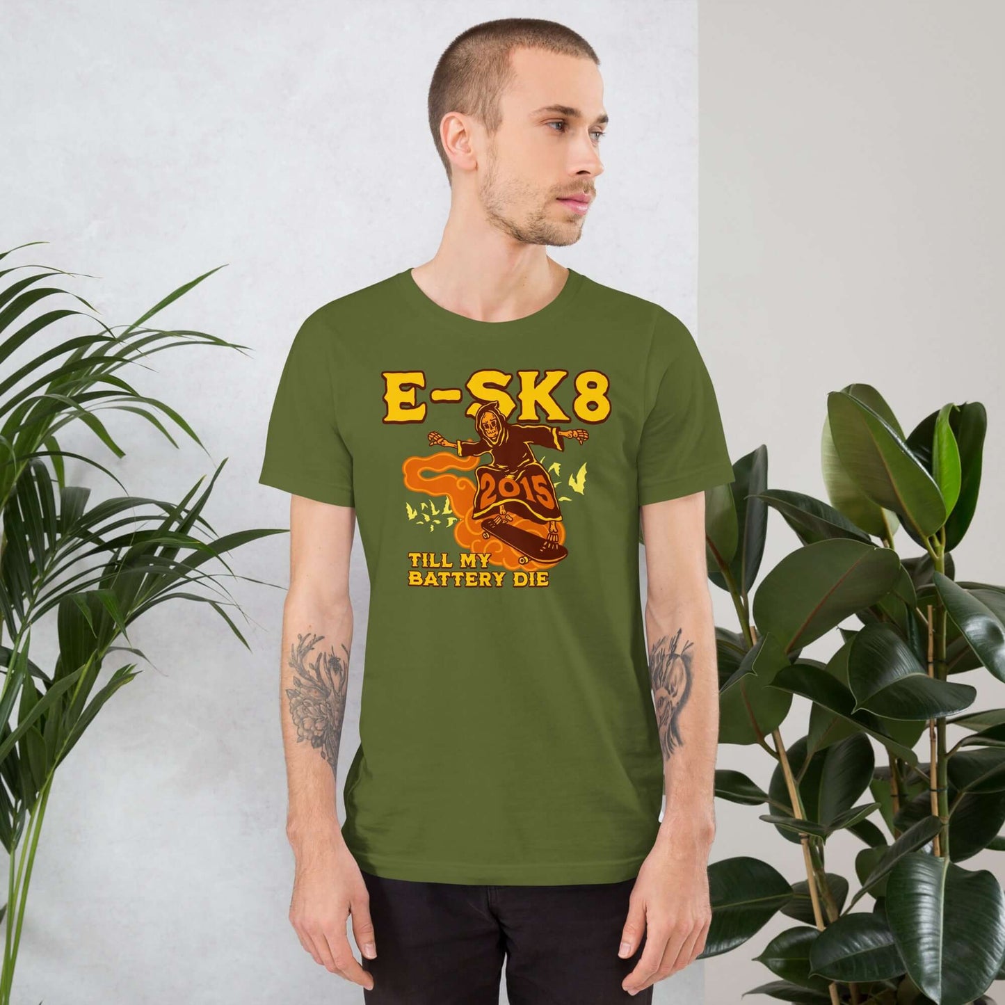 Man wearing E-SK8 Till My Battery Dies T-Shirt with Grim Reaper design in green, standing indoors next to potted plants.