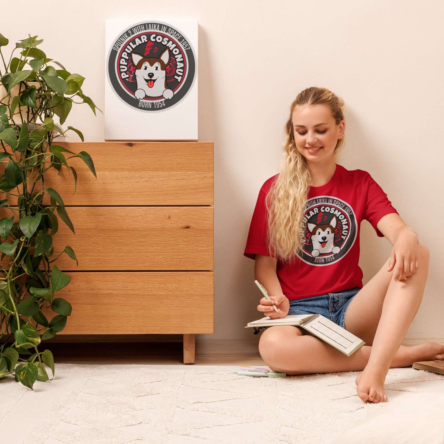 Woman wearing a red "Puppular Cosmonaut" T-shirt with Laika, sitting on the floor next to a dresser and art print.