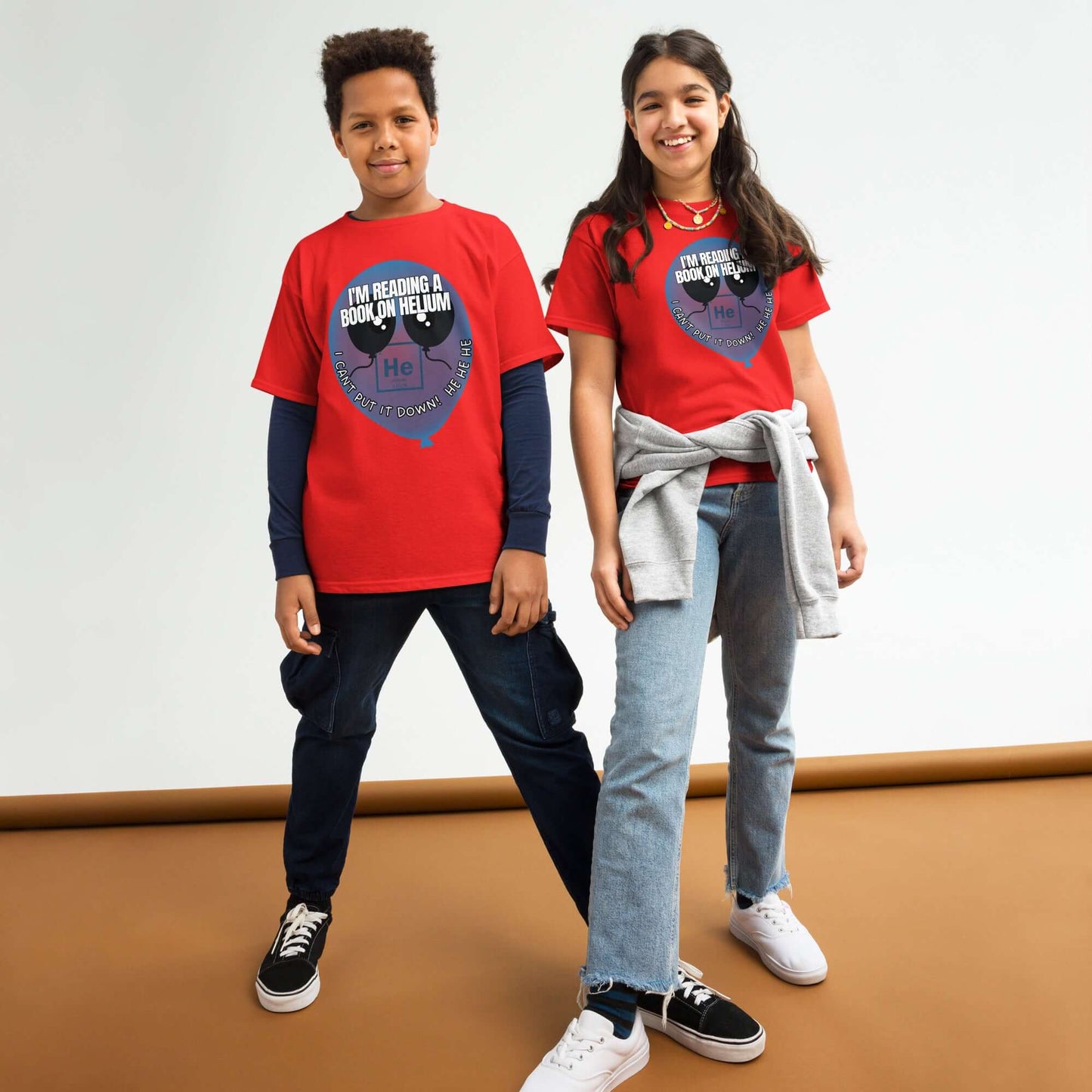 Two children wearing red "I'm Reading a Book on Helium" tees with helium balloon design and chemistry pun, standing in a studio.